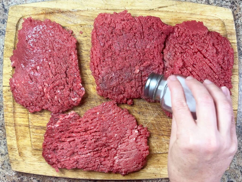 Hand seasoning four cube steaks on a wood cutting board.