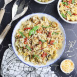 Large serving bowl of bow tie pasta salad with serving spoons and two individual bowls of salad on the side.