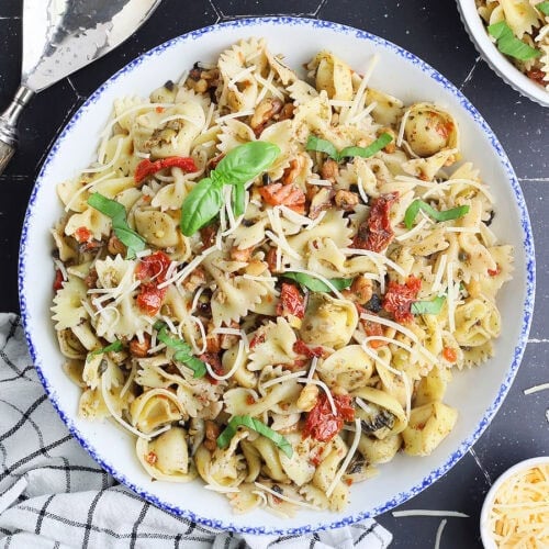 Large serving bowl of bow tie pasta salad with serving spoons and an individual bowl of salad on the side.