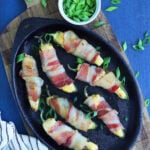 Cast iron plate with seven bacon wrapped jalapeno poppers garnished with sliced green onions.