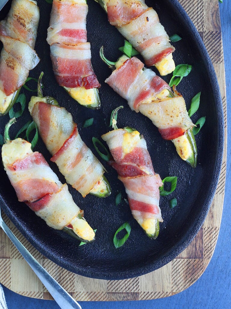 Cast iron plate with seven bacon wrapped jalapeno poppers garnished with sliced green onions.