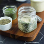 Glass jar of all purpose seasoning on a wooden cutting board with small bowls of herbs and spices in the background.