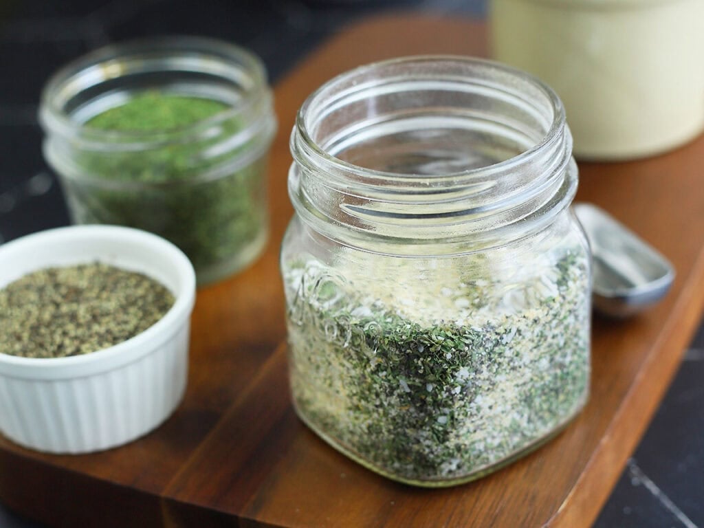 Glass jar of all purpose seasoning on a wooden cutting board with small bowls of herbs and spices in the background.