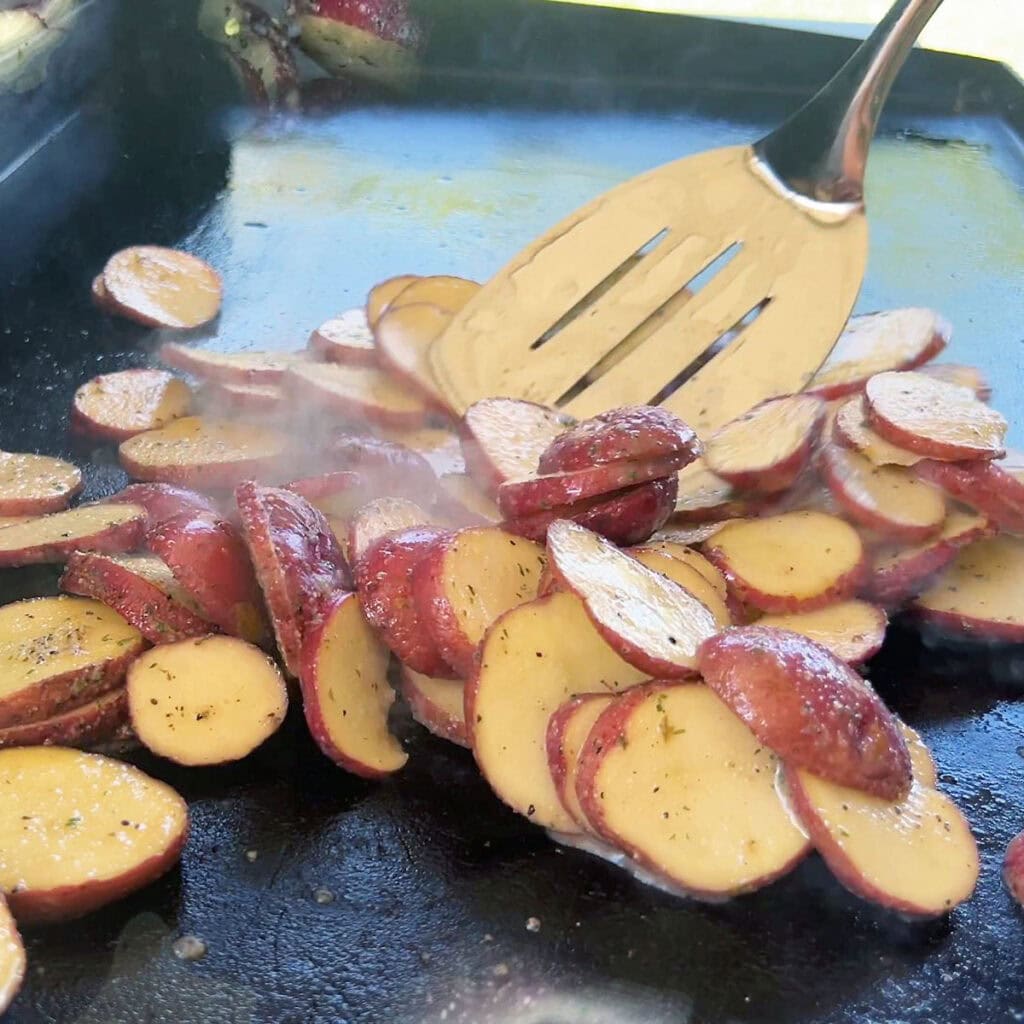 Metal spatula spreading the potato slices over the surface of the hot griddle.
