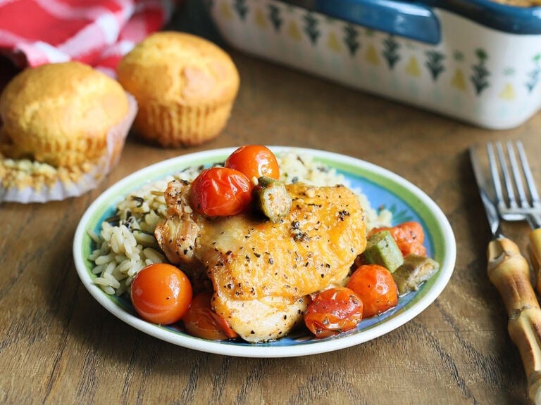 One oven roasted chicken thigh on a plate with rice and topped with okra and tomatoes.