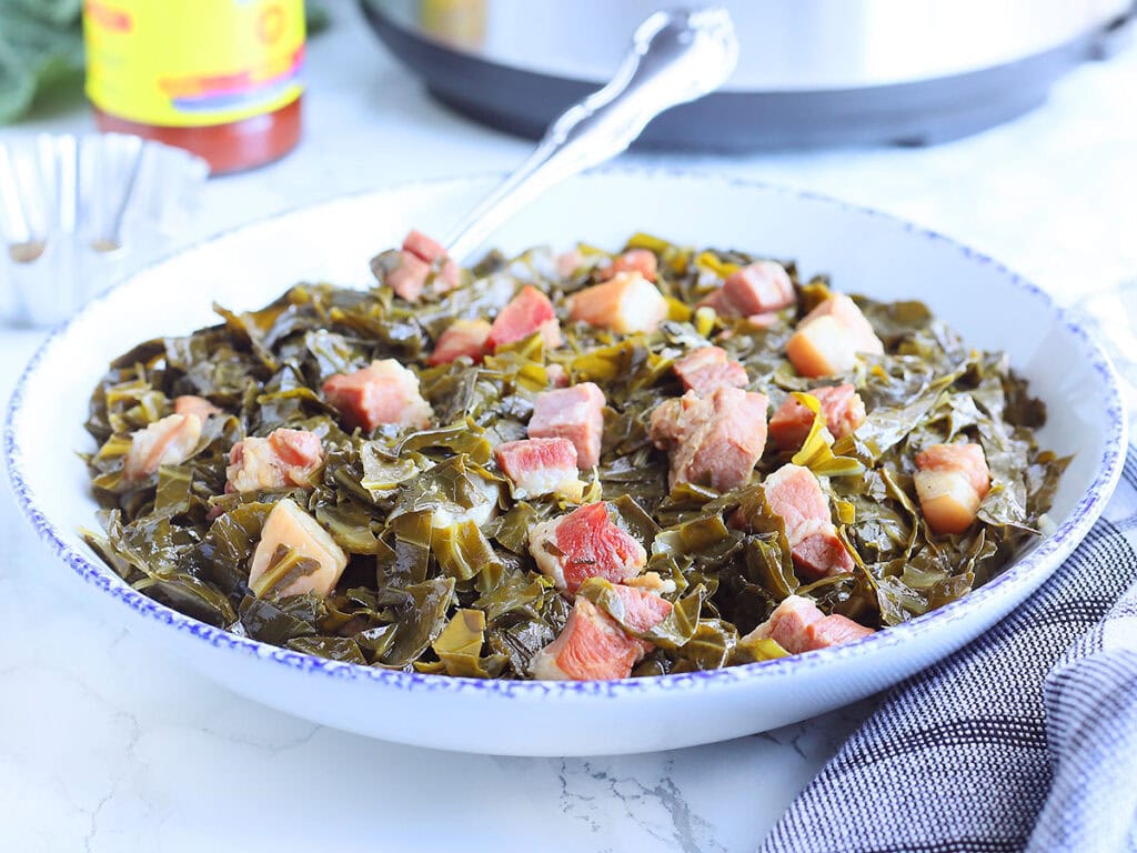 Serving dish full of collard greens with a bottle of hot sauce and an Instant Pot in the background.