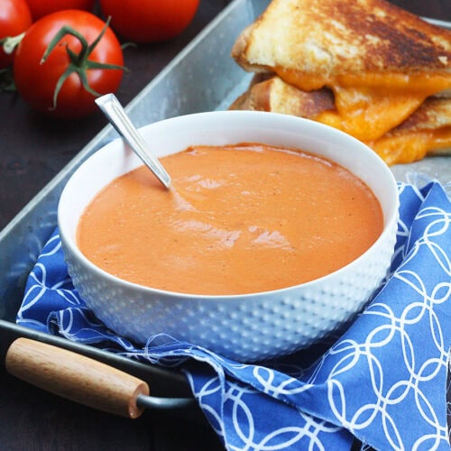 Bowl of tomato basil bisque with a gooey grilled cheese sandwich and fresh tomatoes in the background.