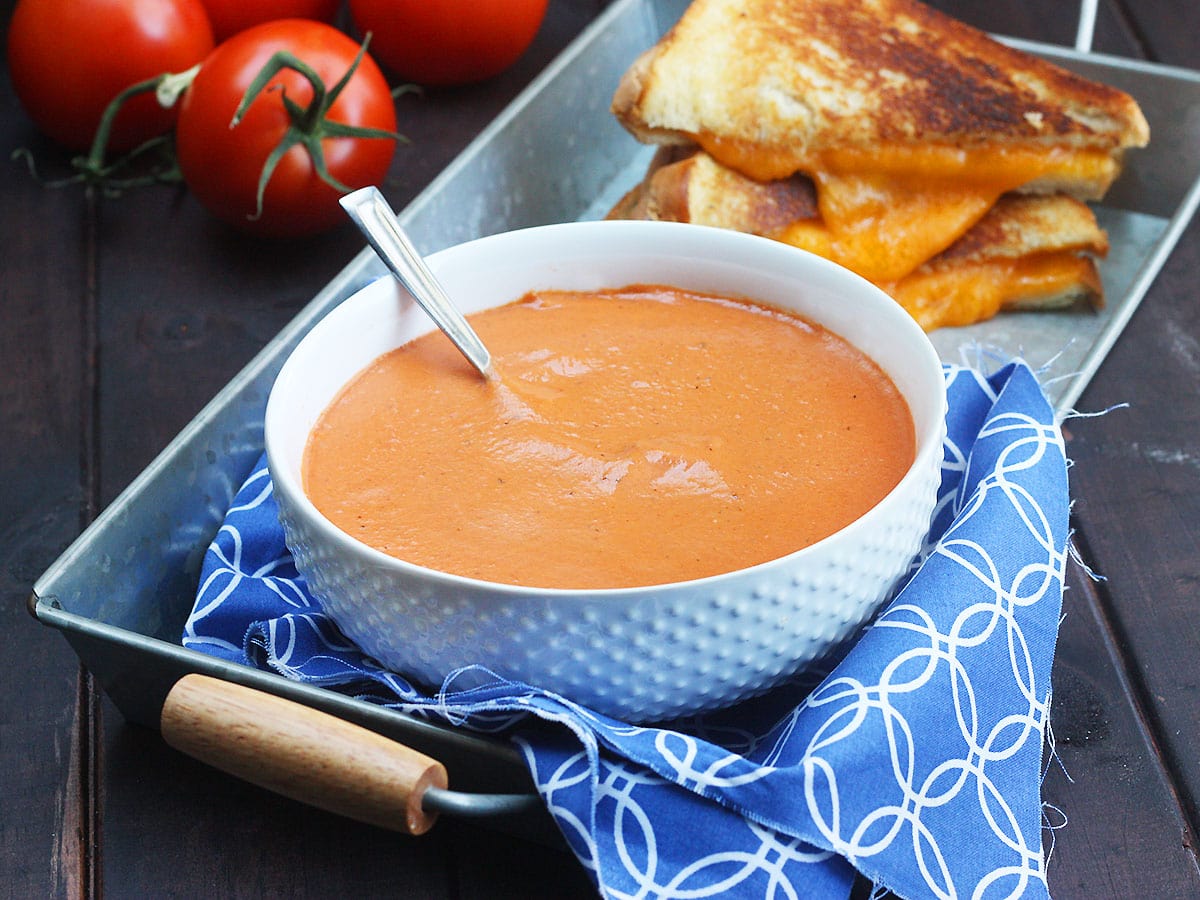 Bowl of tomato basil bisque with a gooey grilled cheese sandwich and fresh tomatoes in the background.