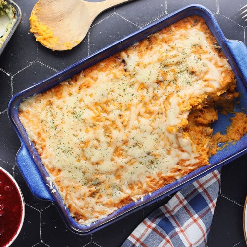 Savory sweet potato casserole with a serving removed. Cranberry sauce, green bean casserole, and a platter of sliced turkey on the side.