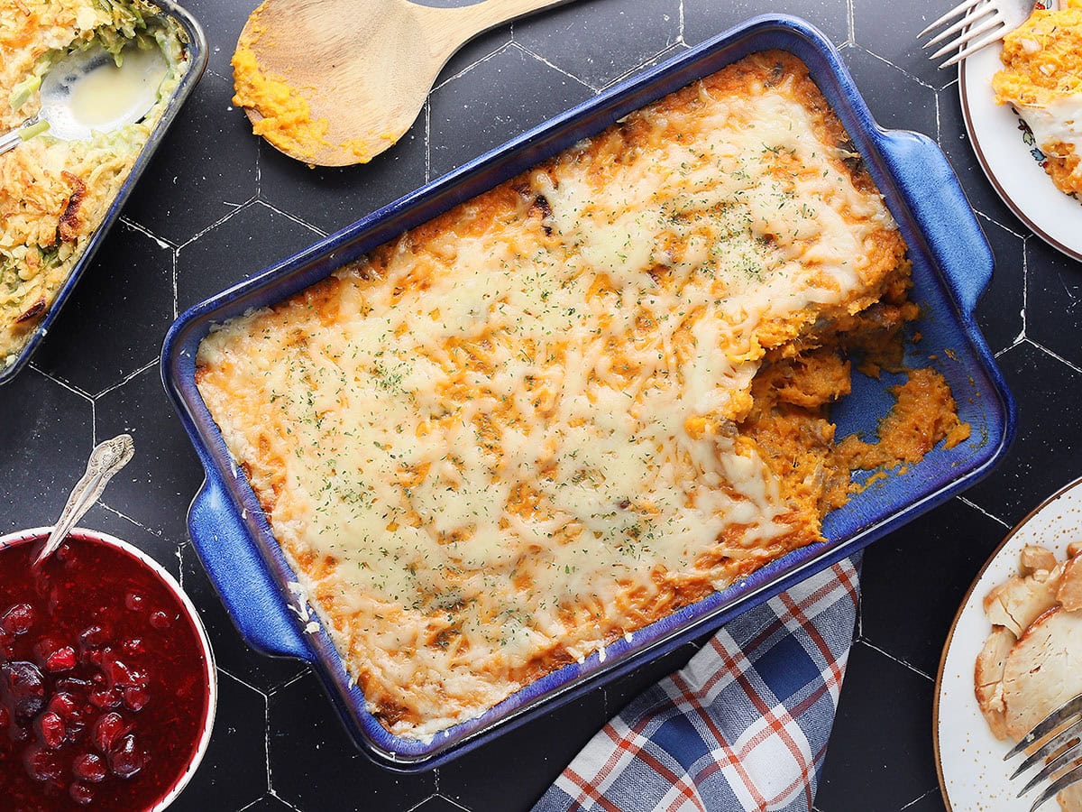 Savory sweet potato casserole with a serving removed. Cranberry sauce, green bean casserole, and a platter of sliced turkey on the side.