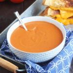 Bowl of tomato basil bisque with a gooey grilled cheese sandwich in the background.