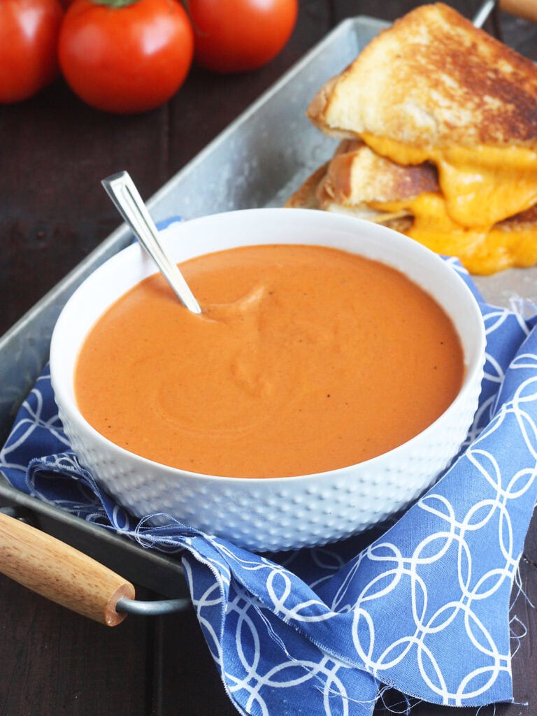 Bowl of tomato basil bisque with a gooey grilled cheese sandwich in the background.