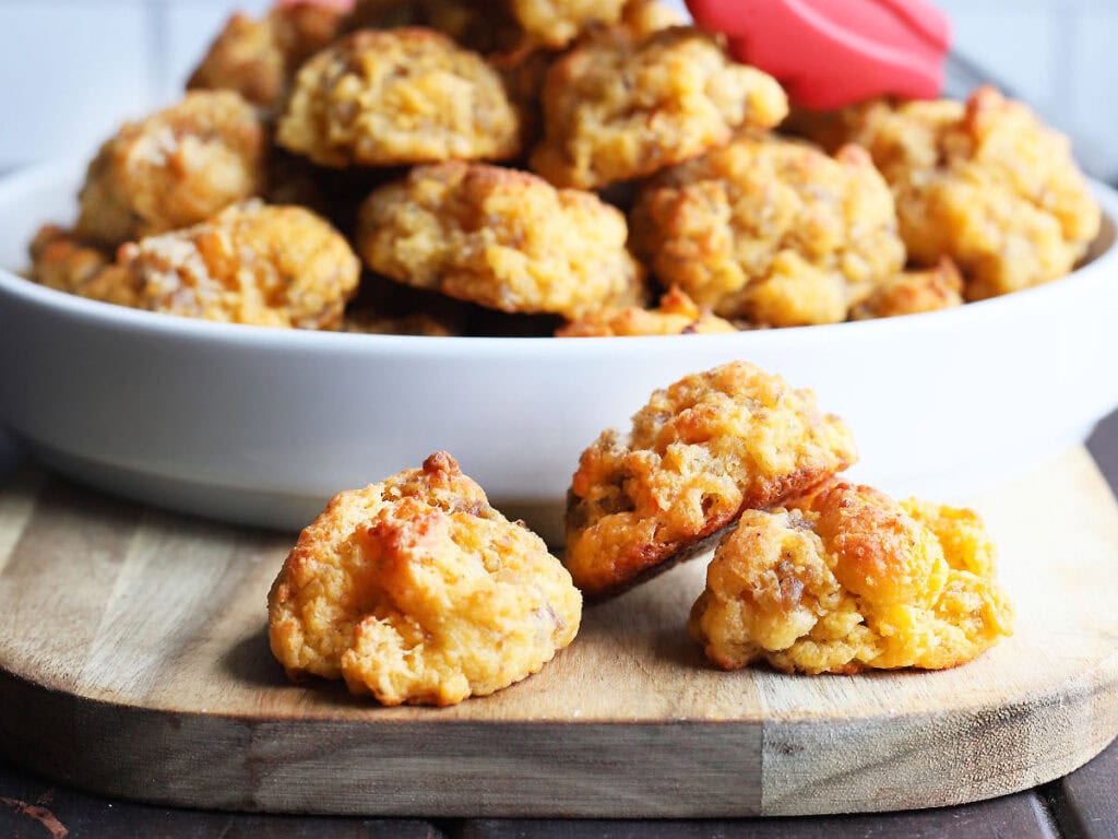 Three sausage balls on a serving board with a serving bowl of sausage balls in the background.