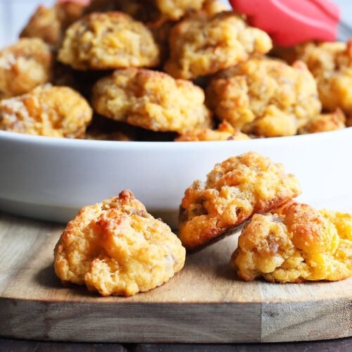 Three sausage balls on a serving board with a serving bowl of sausage balls in the background.