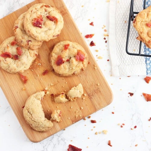 Four bacon and butterscotch chip cookies on a small cutting board, with additional cookies on a cooling rack.
