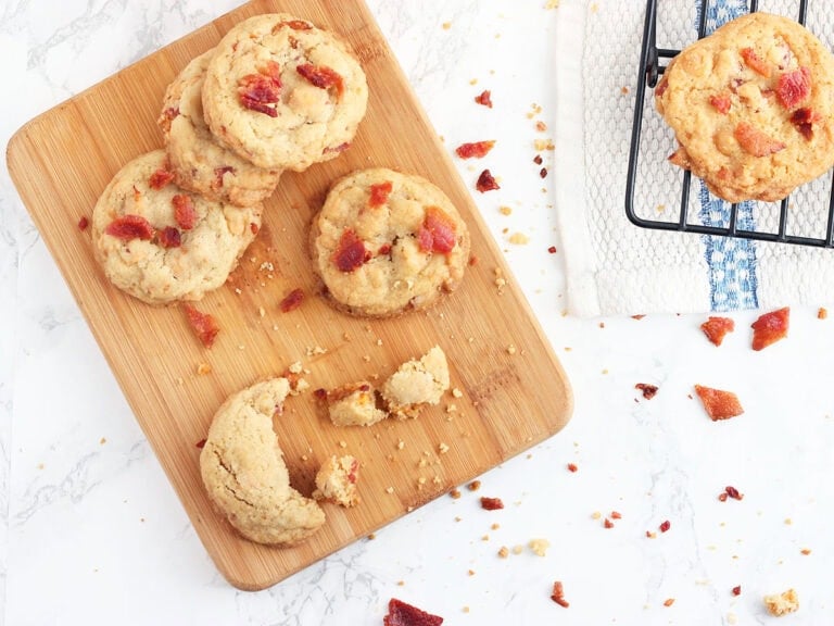 Four bacon and butterscotch chip cookies on a small cutting board, with additional cookies on a cooling rack.