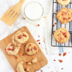 Four bacon and butterscotch chip cookies on a small cutting board, with additional cookies on a cooling rack, a glass of milk and a wooden spatula on the side.