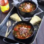 Two bowls of vegetable soup with collard greens garnished with shredded ham and served with wedges of cornbread on a metal tray.