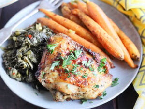 Single air fryer lemon pepper chicken thigh on a plate with collard greens and roasted carrots.