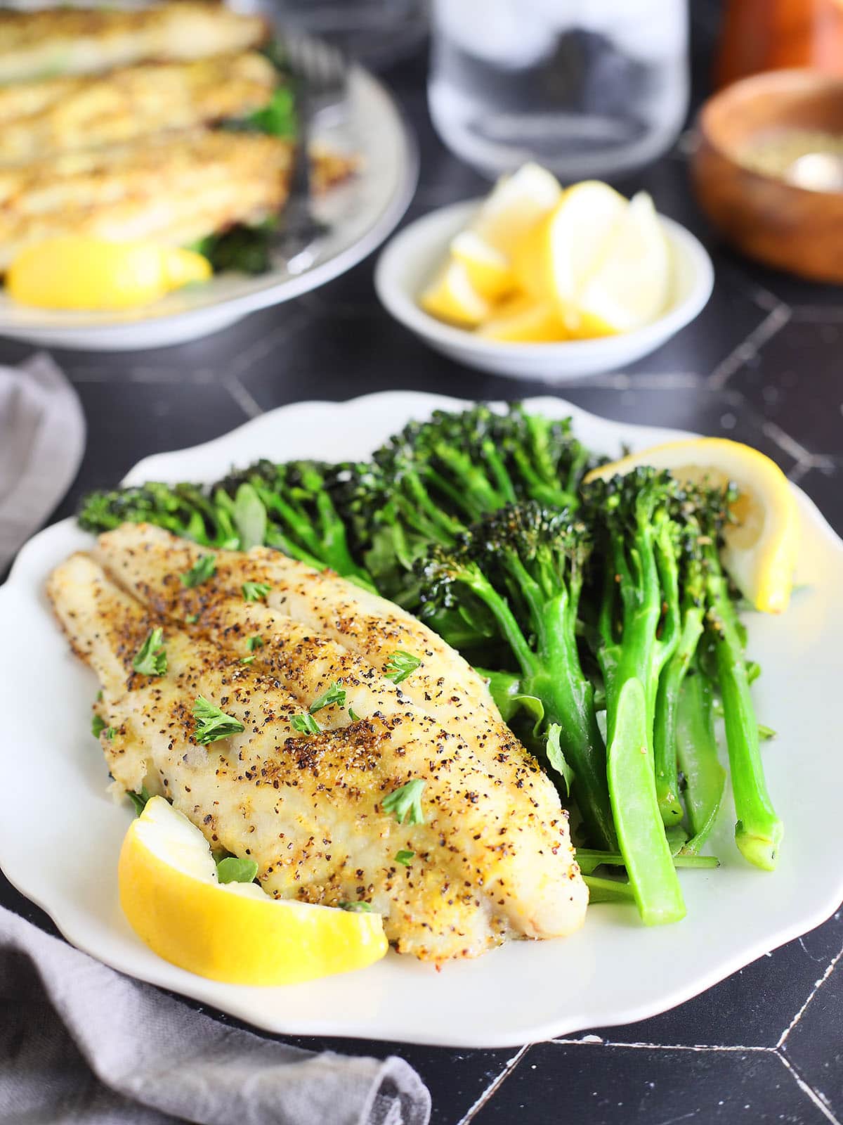 Lemon pepper air fryer catfish fillet on a plate with broccolini and lemon wedges.