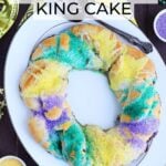 Whole decorated crescent roll king cake on a white platter with Mardi Gras decorations around it.