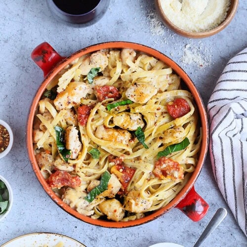 Serving bowl of Marry Me Chicken Pasta with two glasses of red wine, small dishes of grated Parmesan and sundried tomatoes, and an individual plate of pasta on the side.