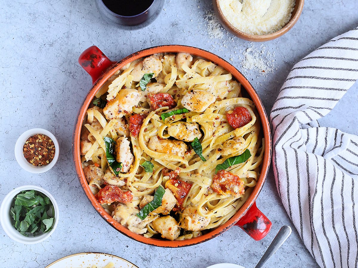 Serving bowl of Marry Me Chicken Pasta with two glasses of red wine, small dishes of grated Parmesan and sundried tomatoes, and an individual plate of pasta on the side.