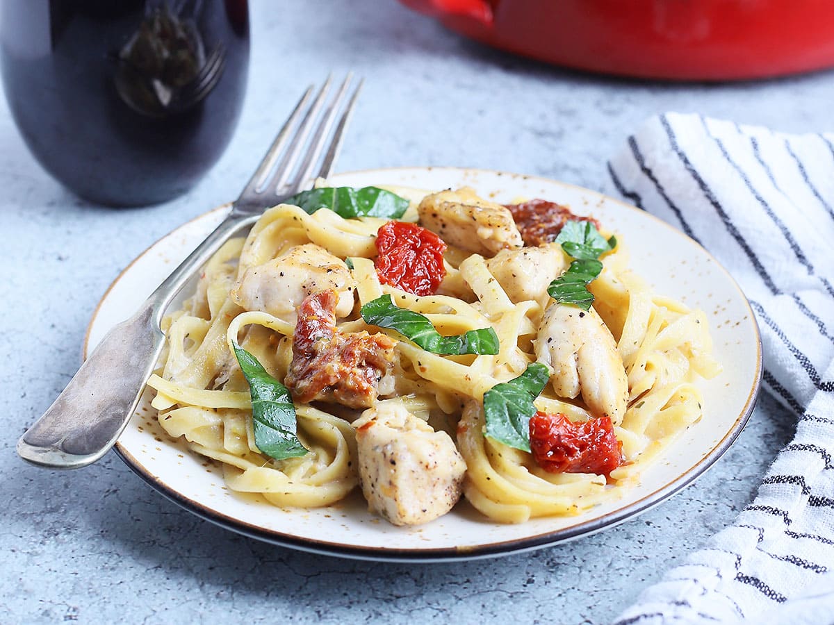 Serving of Marry Me Chicken Pasta with a glass of red wine and a serving bowl of pasta in the background.