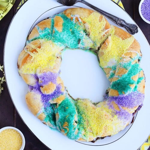 Whole decorated crescent roll king cake on a white platter with Mardi Gras decorations around it.