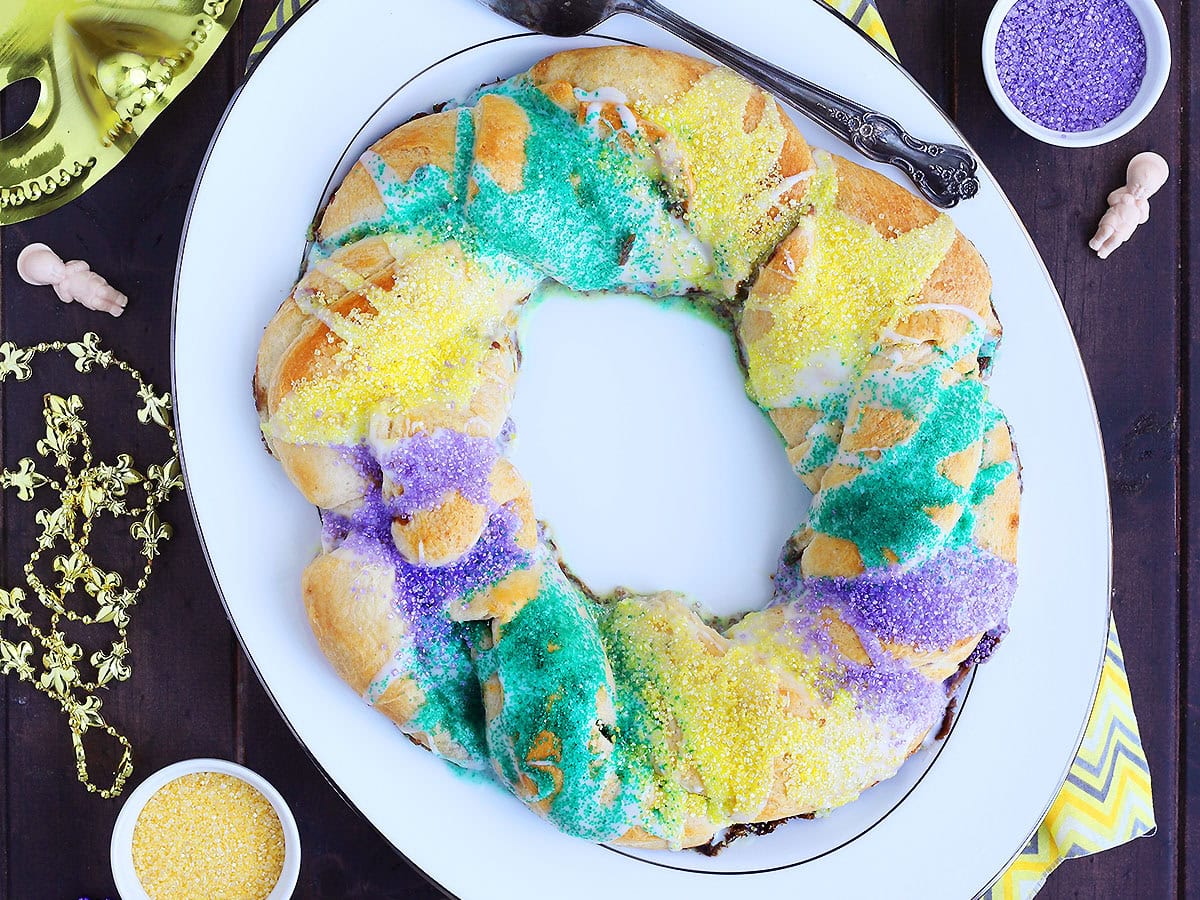 Whole decorated crescent roll king cake on a white platter with Mardi Gras decorations around it.