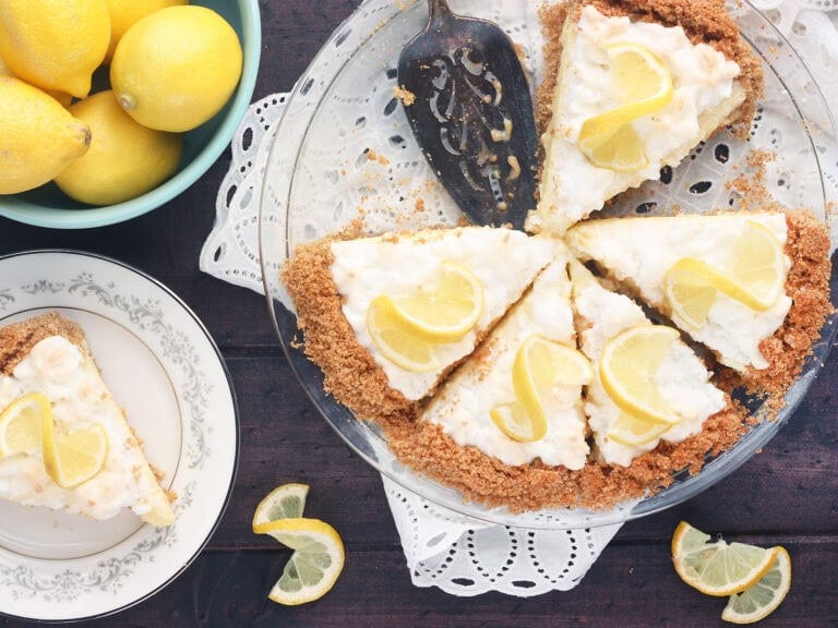 Sliced lemon icebox pie with a single serving of pie to the side on a plate and a bowl of fresh lemons to the side.