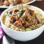 Hand pouring teriyaki sauce over a bowl of bourbon chicken served over rice.