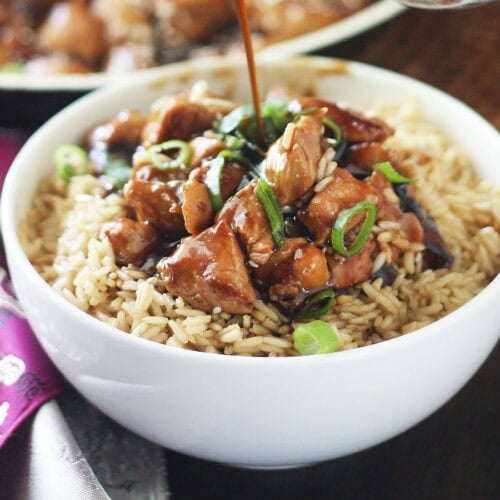 Hand pouring teriyaki sauce over a bowl of bourbon chicken served over rice.