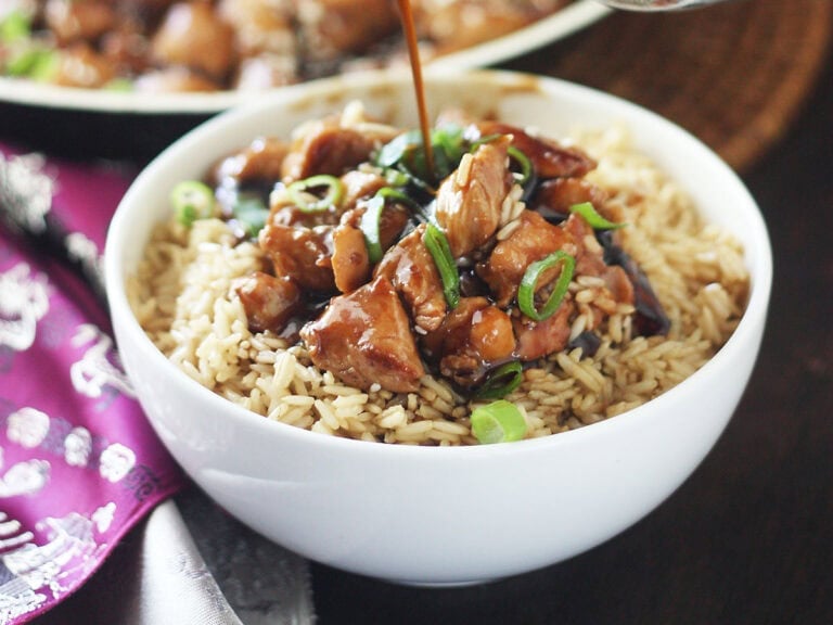 Hand pouring teriyaki sauce over a bowl of bourbon chicken served over rice.