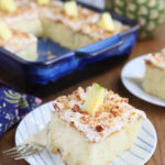 Single slice of pineapple poke cake on a plate with the rest of the cut cake and a whole pineapple in the background.