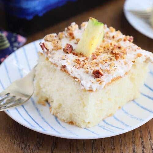 Single slice of pineapple poke cake on a plate with the rest of the cut cake and a whole pineapple in the background.