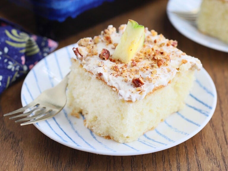 Single slice of pineapple poke cake on a plate with the rest of the cut cake and a whole pineapple in the background.