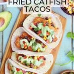 Four catfish tacos garnished with salsa and avocado crema sauce on a wooden cutting board.