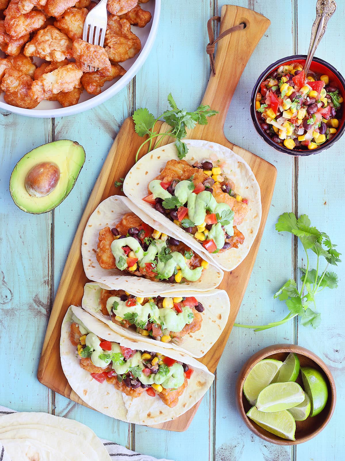 Four catfish tacos garnished with salsa and avocado crema sauce on a wooden cutting board.