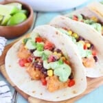 One catfish taco garnished with avocado cream sauce and salsa on a wooden cutting board.