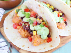 One catfish taco garnished with avocado cream sauce and salsa on a wooden cutting board.