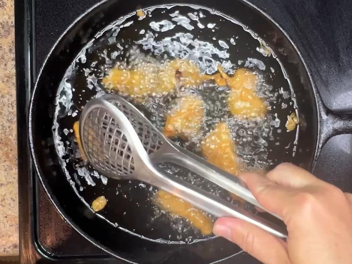Hand using metal tongs to flip catfish pieces in hot oil.