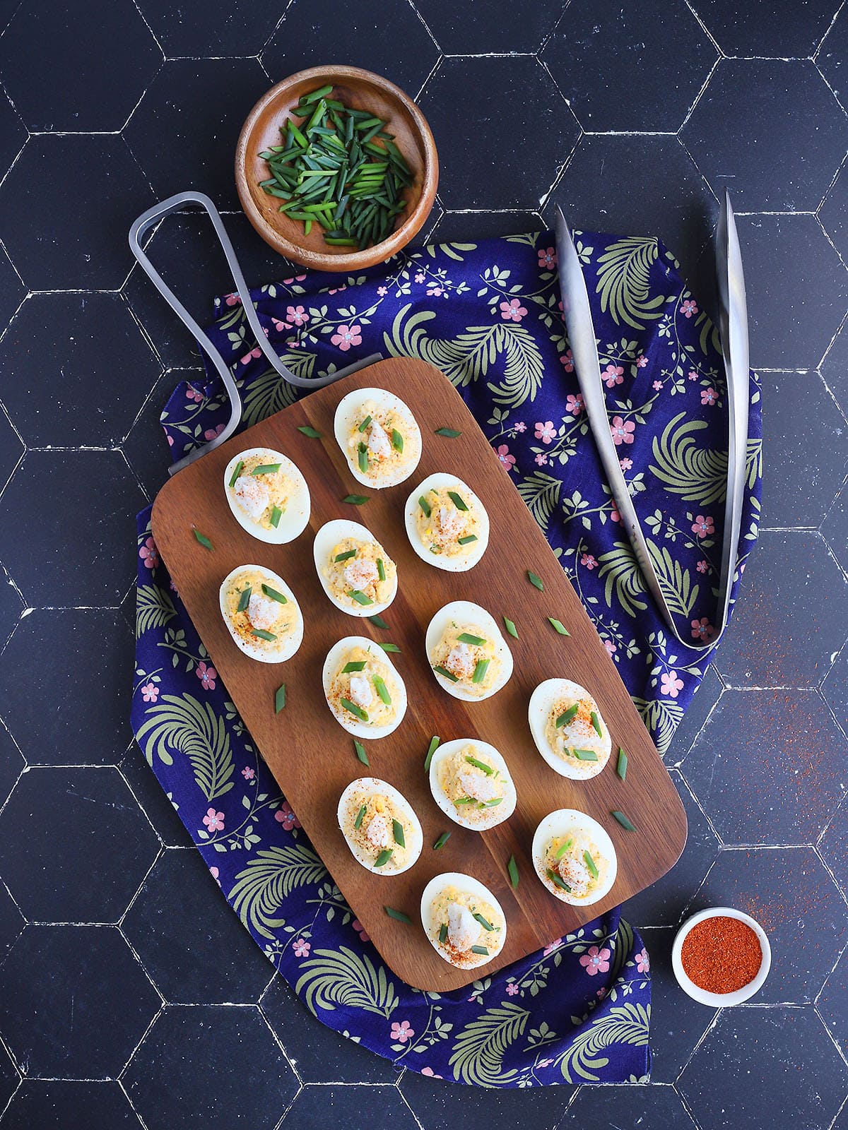 Crab deviled eggs garnished with crab meat, paprika and fresh chives on a wooden cutting board with metal serving tongs, a small bowl of chopped fresh chives and a dish of paprika on the side.