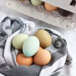 Multicolored eggs wrapped in a hand towel with a carton of a dozen eggs in the background.