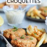 Two fried salmon croquettes garnished with fresh parsley on a plate with a fork and some French fries on the side. A dish of tartar sauce, a second plate of food and a platter of cooked salmon croquettes can be seen in the background.