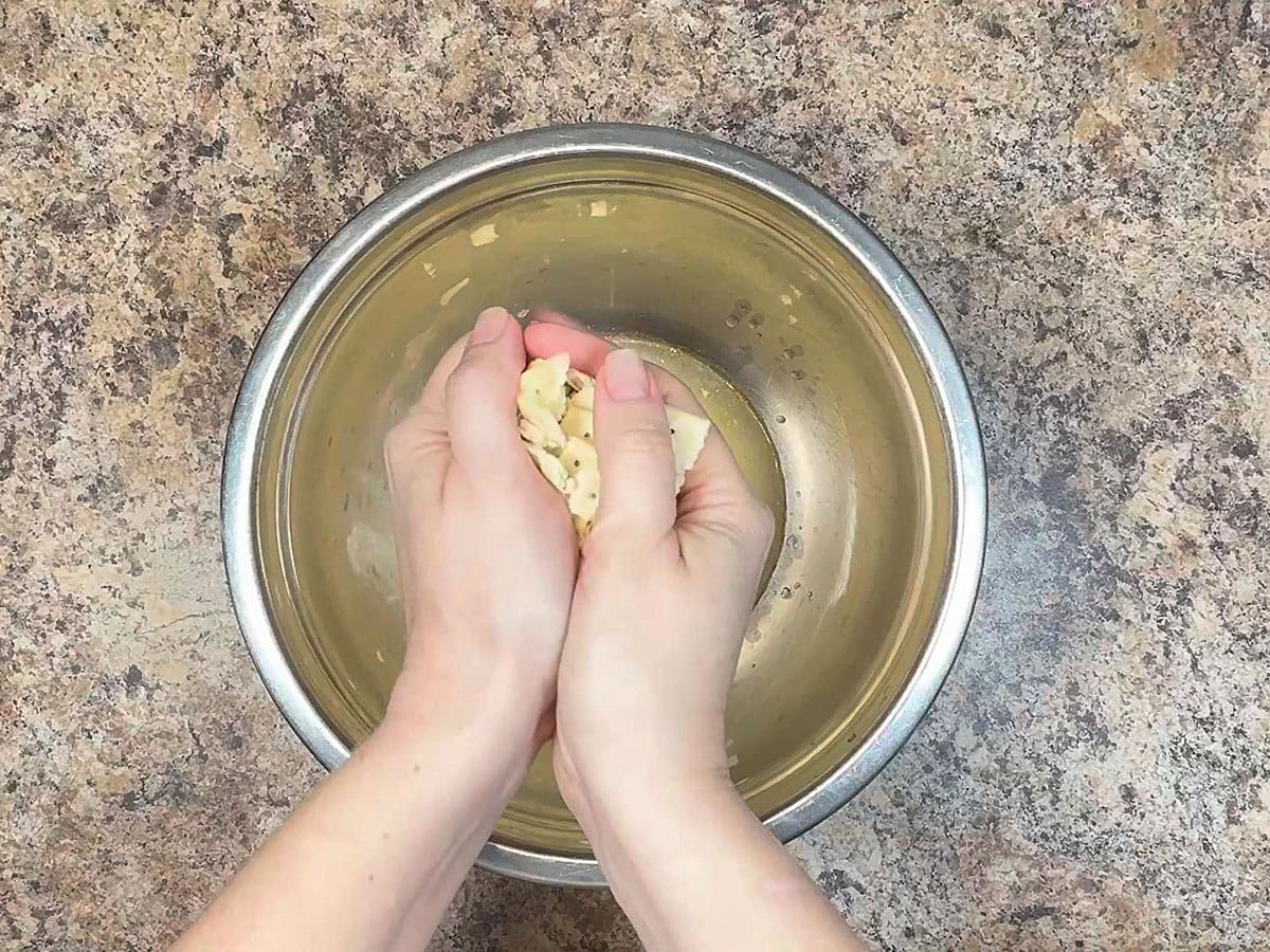 Pair of hands crushing saltine crackers over a mixing bowl of liquid drained from the can of salmon.