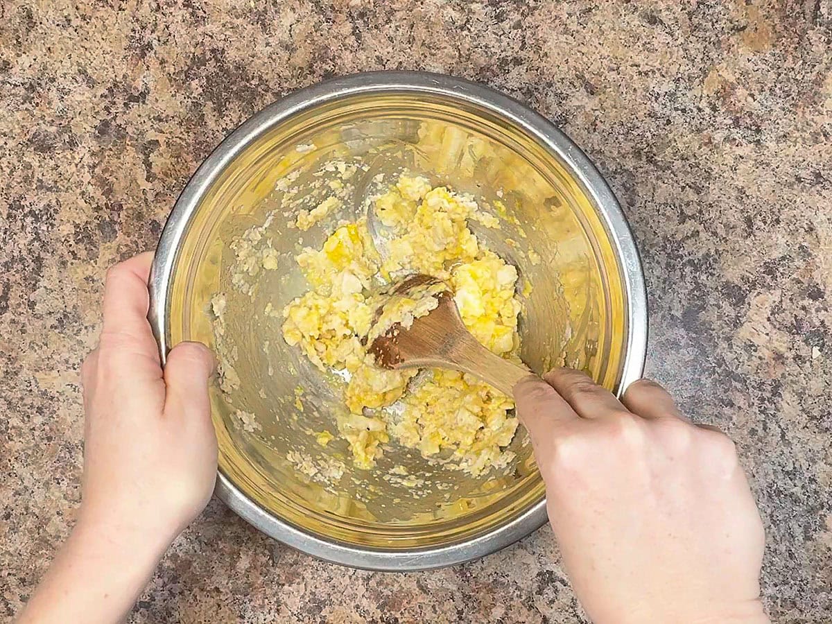 Hands using a wooden spoon to combine the egg with the crushed crackers in a large mixing bowl.