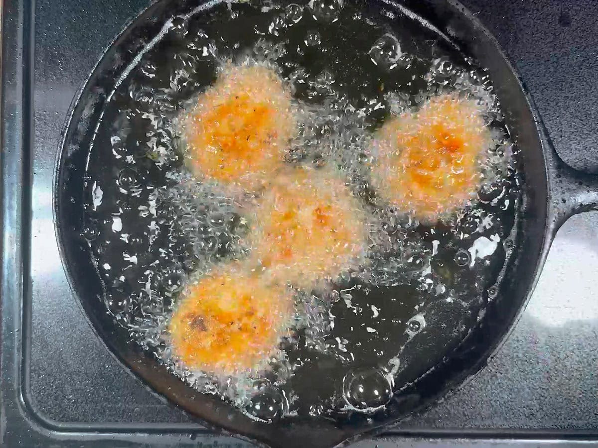 Four salmon croquettes frying in a cast iron skillet on a stove.