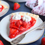 Slice of strawberry pie with jello topped with whipped cream on a plate with a fork on the side.