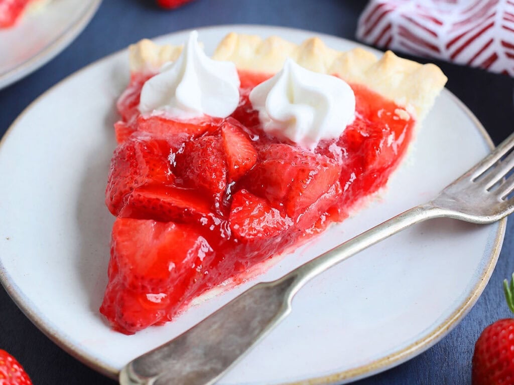 Slice of strawberry pie with jello topped with whipped cream on a plate with a fork on the side.